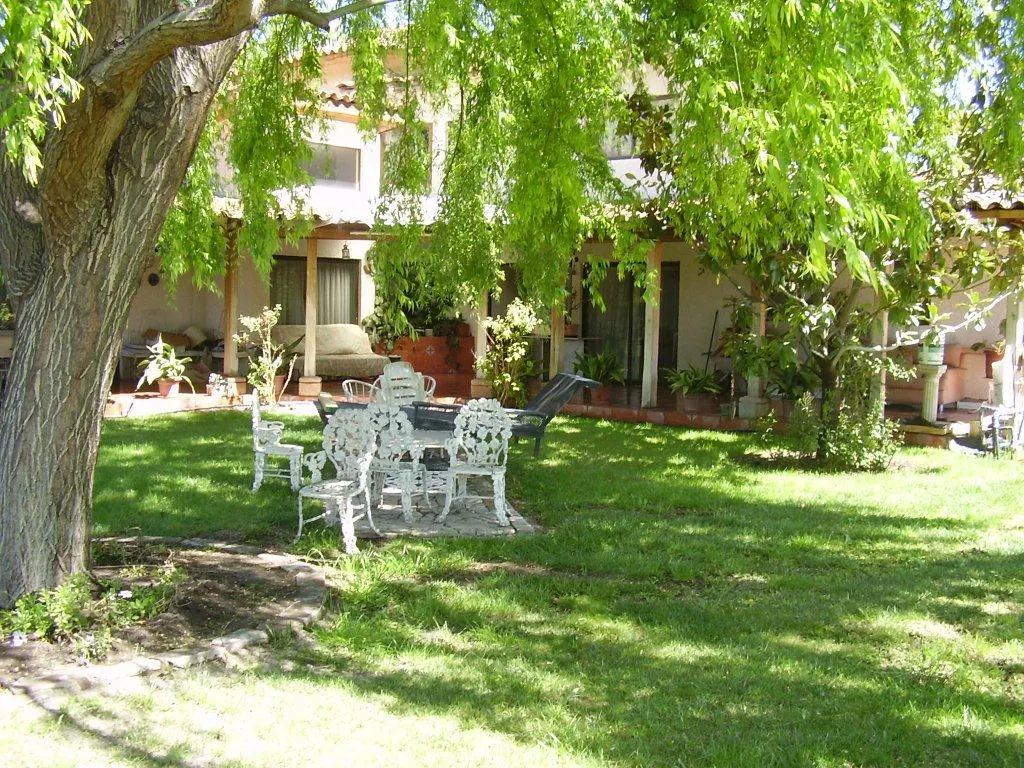 A lush green backyard with a white metal table and chairs set under a large tree. The house has a covered patio with outdoor furniture.