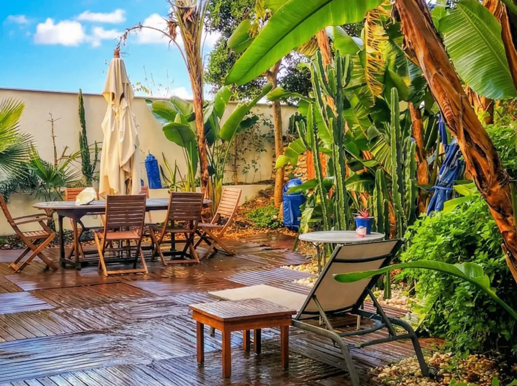 Outdoor patio with wooden deck, table, chairs, lounge chair, and lush tropical plants under a blue sky.
