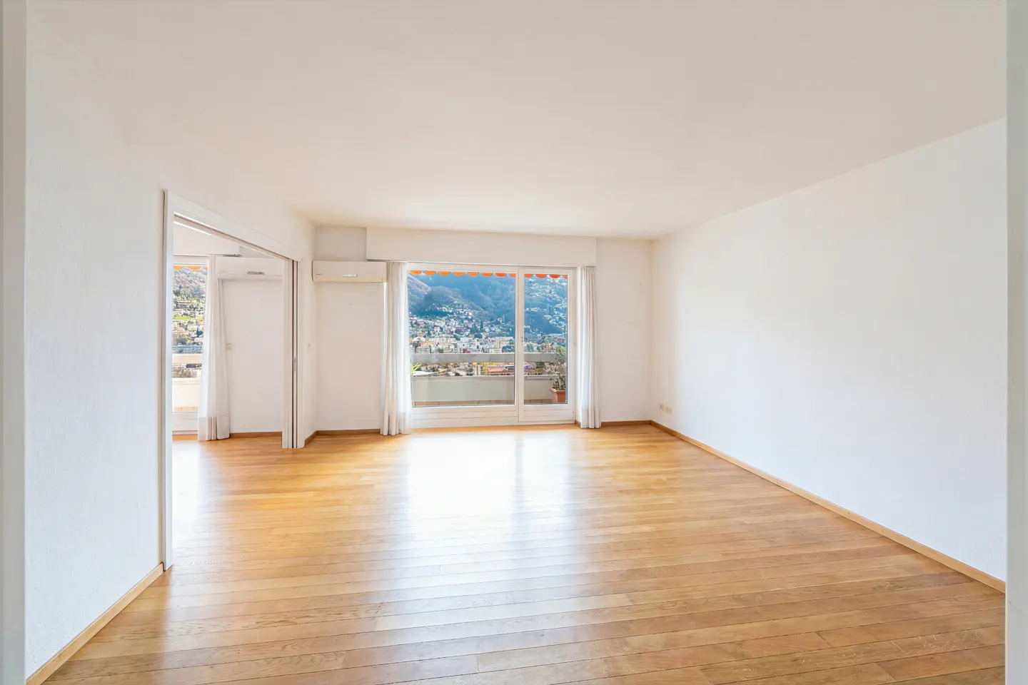 Bright, empty room with hardwood floors and white walls. Large windows offer a view of a city and mountains.