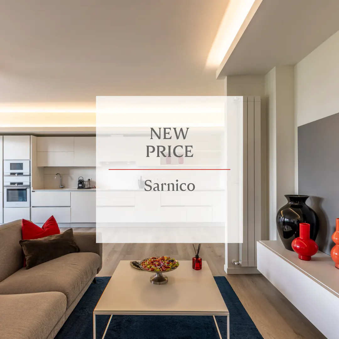 Bright, modern living room with a gray sofa, white kitchen, and a "New Price Sarnico" overlay. A white coffee table sits on a blue rug.