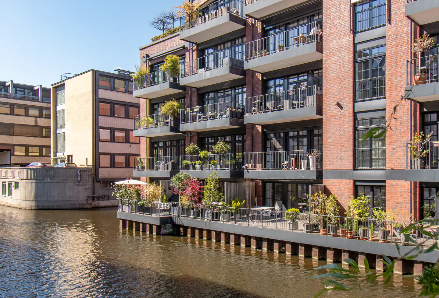 Brick apartment building with black-framed windows and balconies overlooking a canal on a sunny day.