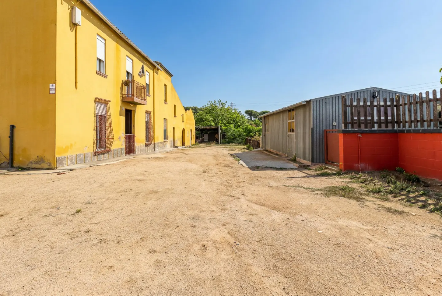 Exterior view of a yellow building with a balcony, next to a metal shed with a red base, under a clear blue sky.