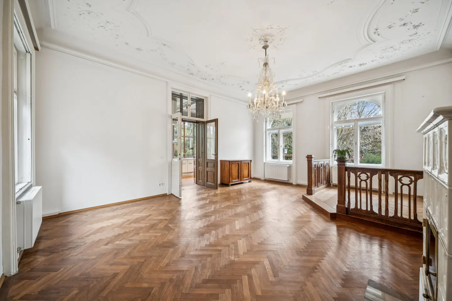 Bright, empty room with herringbone wood floors, white walls, and a crystal chandelier. Windows and an open door let in natural light.