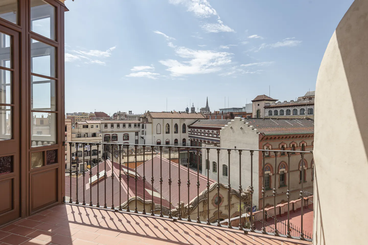 View from a balcony with a black railing overlooking a European city with red-tiled roofs under a blue sky. Brown door on the left.