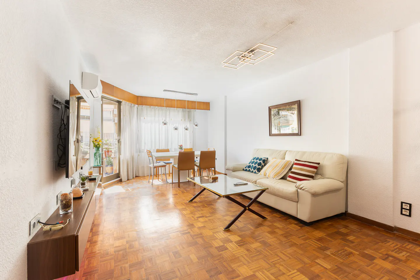 Bright living room with parquet floors, white walls, a beige sofa, and a dining table near a window.