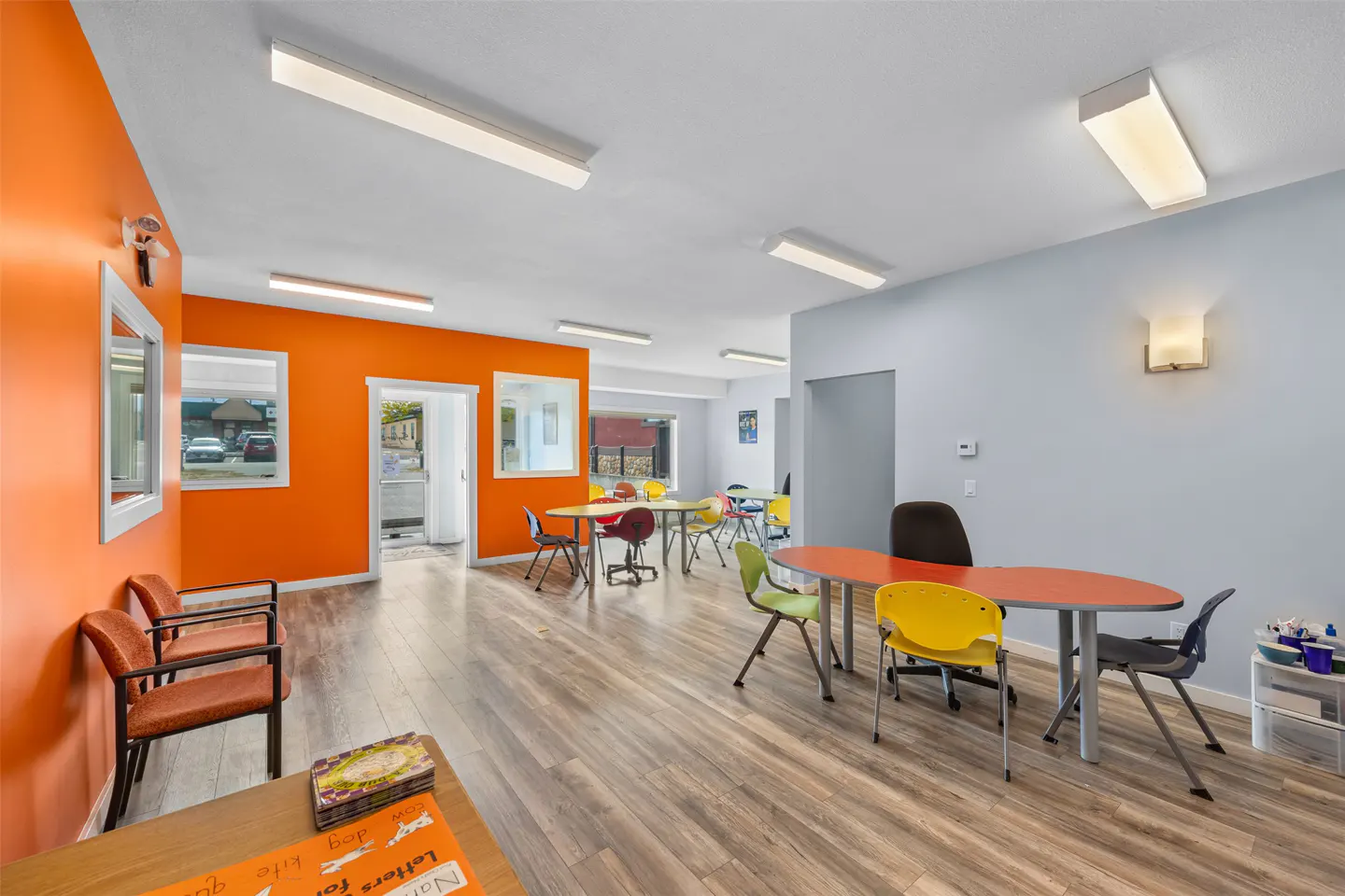 Bright room with orange and gray walls, wood floors, and colorful tables and chairs. Fluorescent lights illuminate the space.
