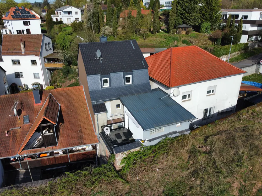 Aerial view of houses with red and gray roofs, surrounded by trees and greenery. A patio with furniture is visible.