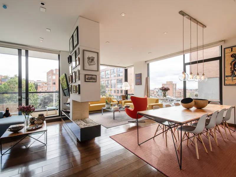 Bright, modern living room with wood floors, large windows, and city views. Yellow sofas, red chair, and dining table with pendant lights.