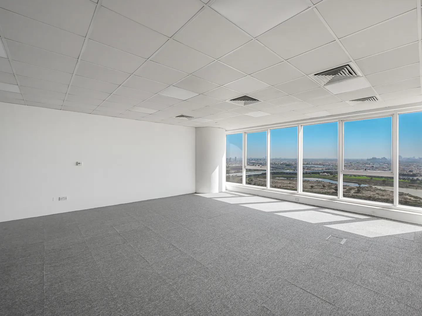 Empty office space with gray carpet, white walls, and a large window showing a city skyline under a blue sky.