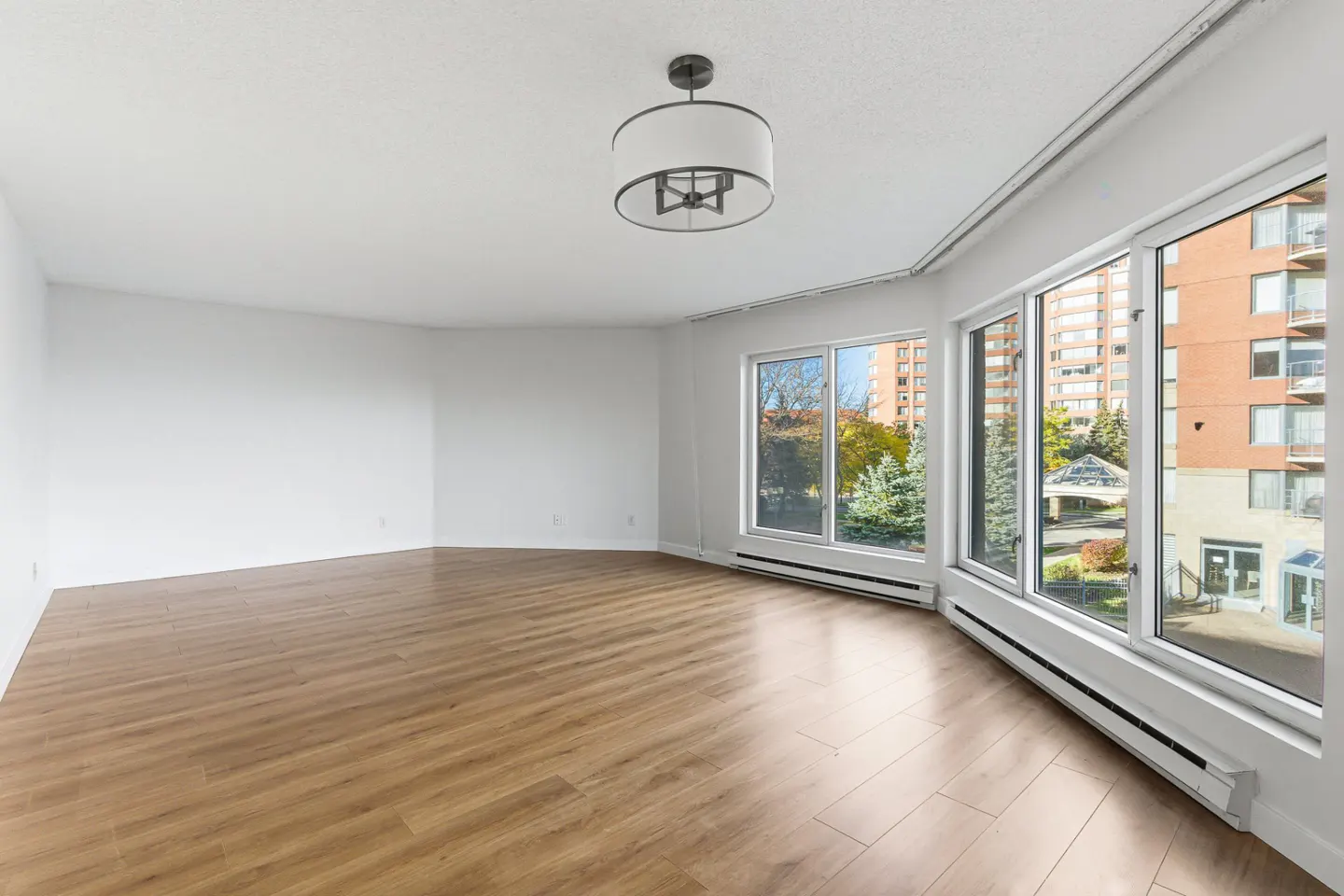 Empty room with wood floors, white walls, and large windows showing a building and trees. A modern light fixture hangs from the ceiling.