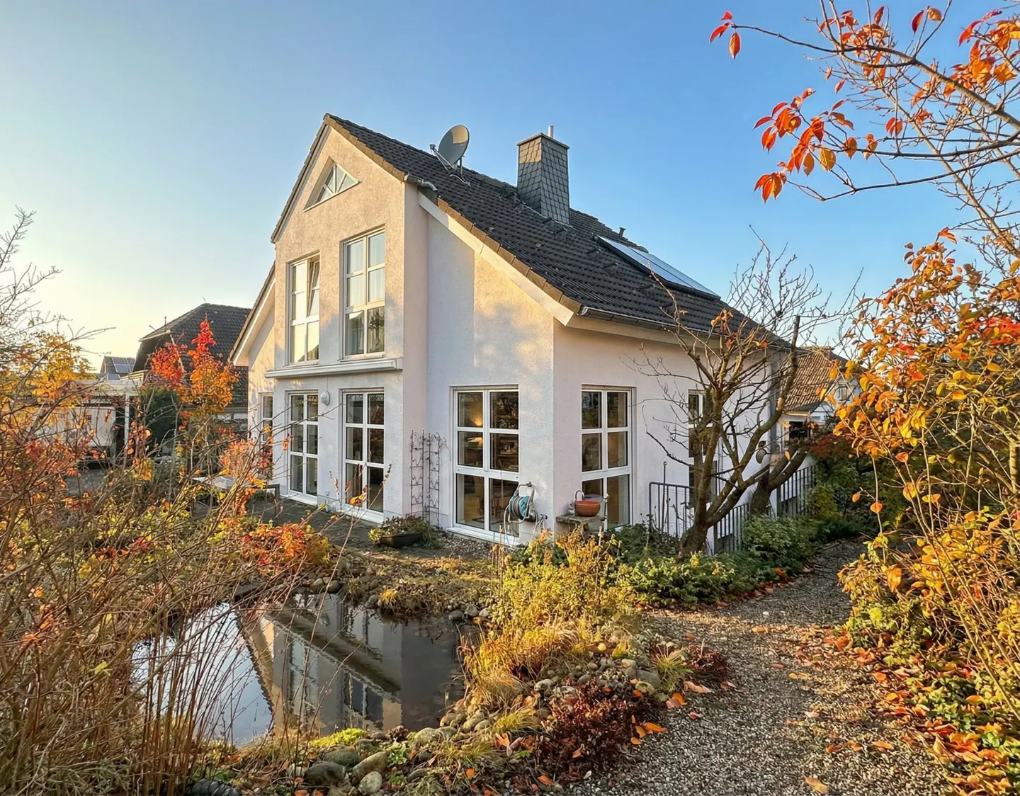 Two-story house with white walls, gray roof, and many windows, surrounded by a garden with a pond and colorful autumn foliage.