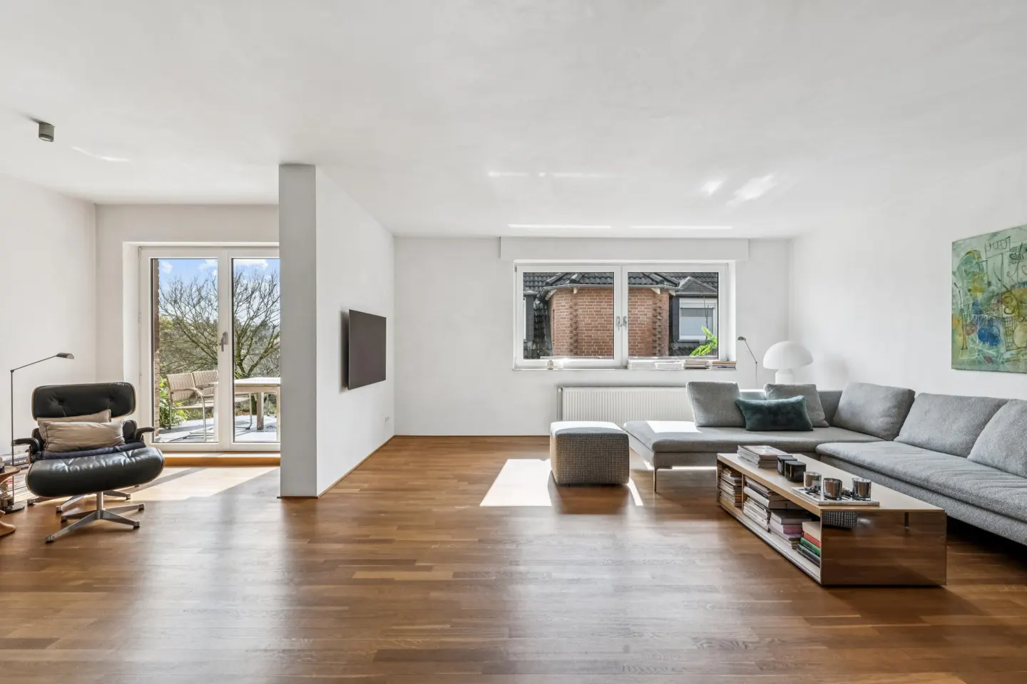 Bright living room with hardwood floors, white walls, and a gray sectional sofa. A black chair sits near a glass door to a balcony.