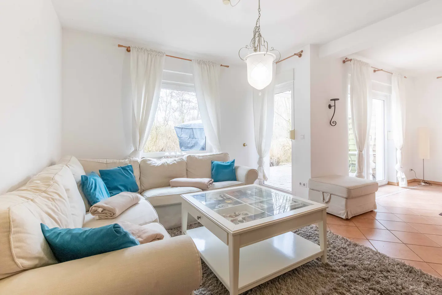Bright living room with a beige sectional sofa, blue pillows, and a white coffee table on a gray rug. White walls and curtains.