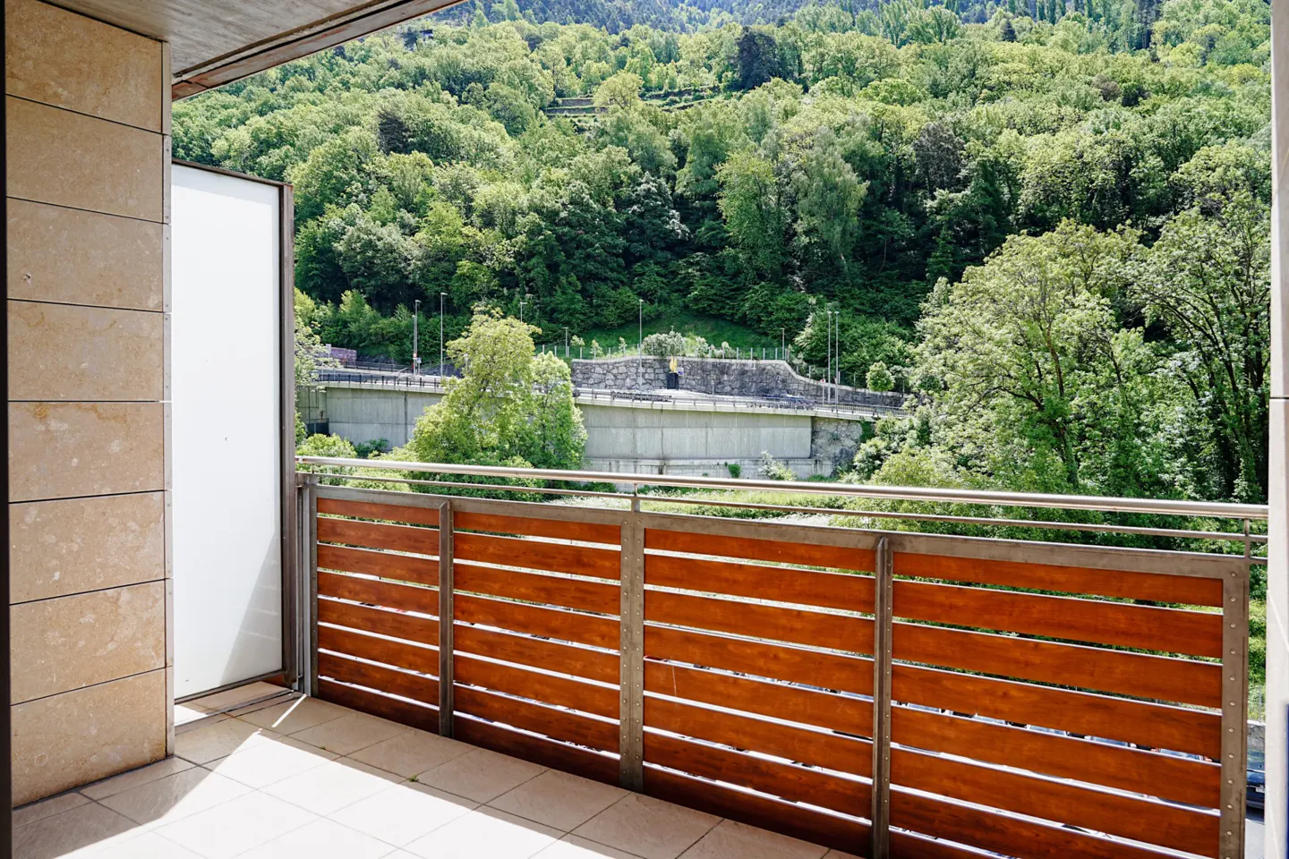 Balcony view with wood railing overlooking lush green trees and a concrete structure in the background.