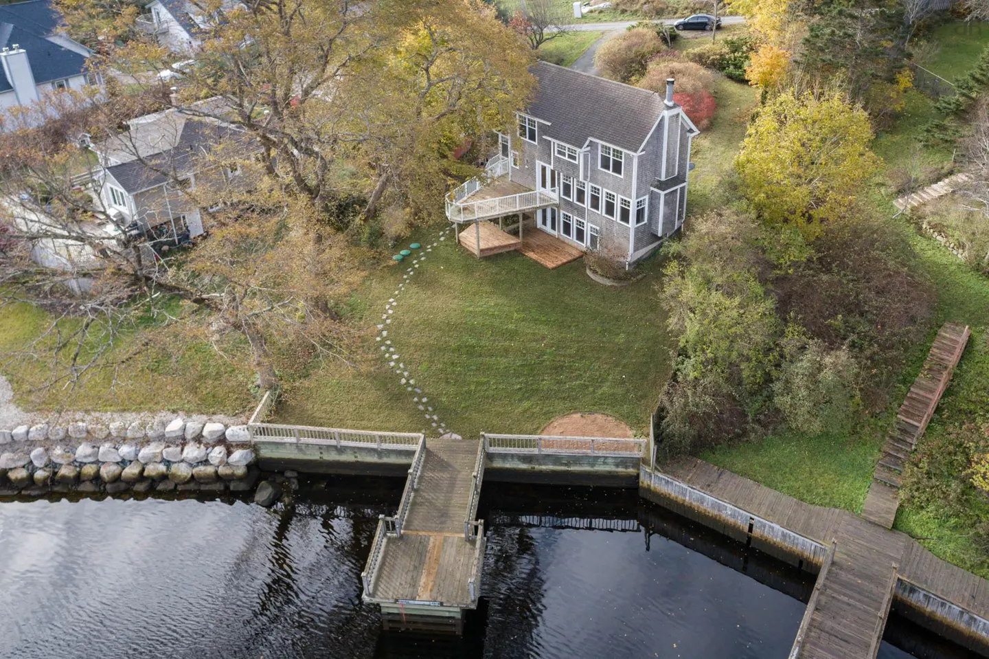Aerial view of a gray two-story house with a deck, a dock on the water, and a stone path in the yard.