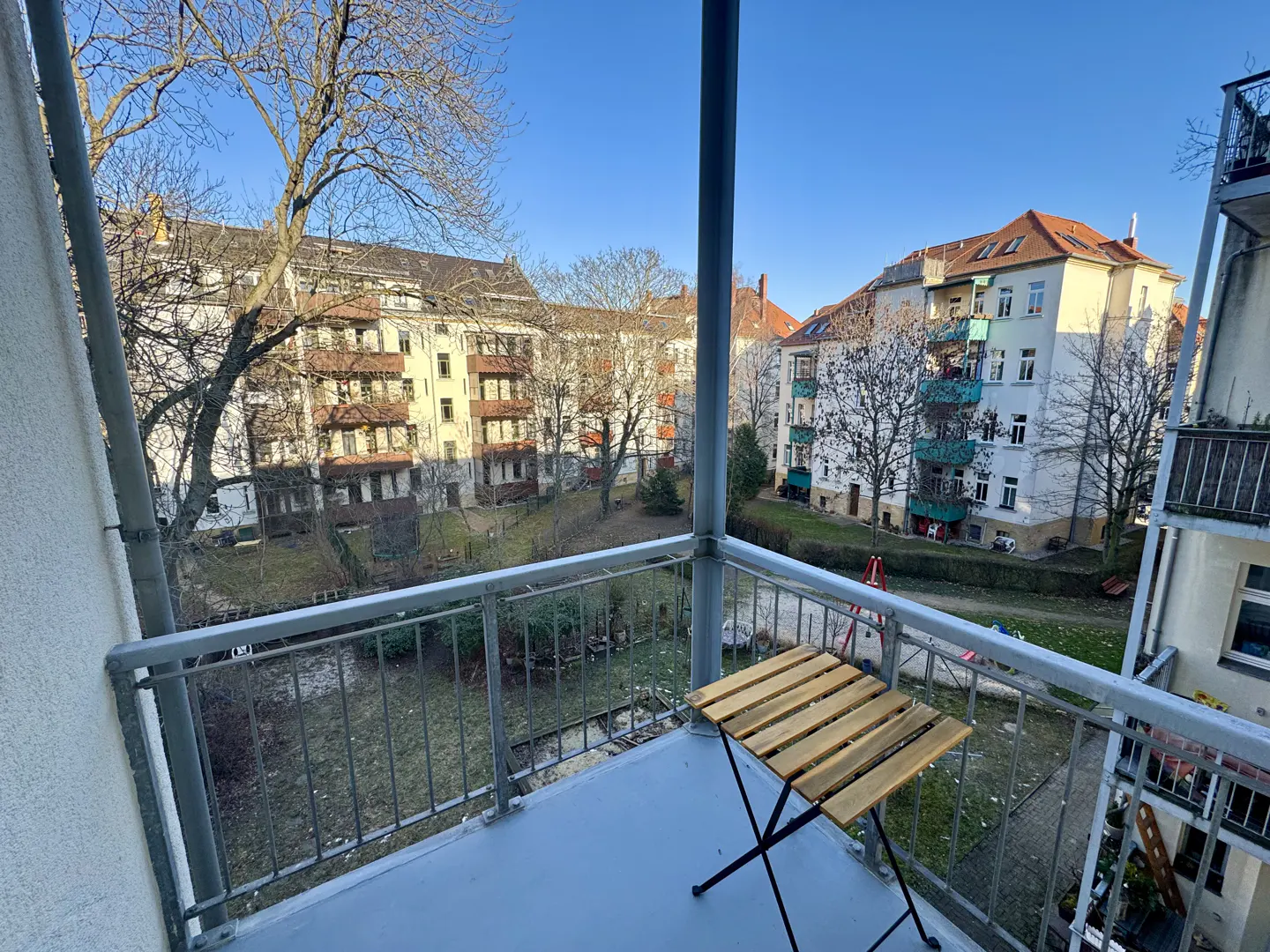 Balcony view with a small wooden table, overlooking a courtyard with buildings and trees under a clear blue sky.