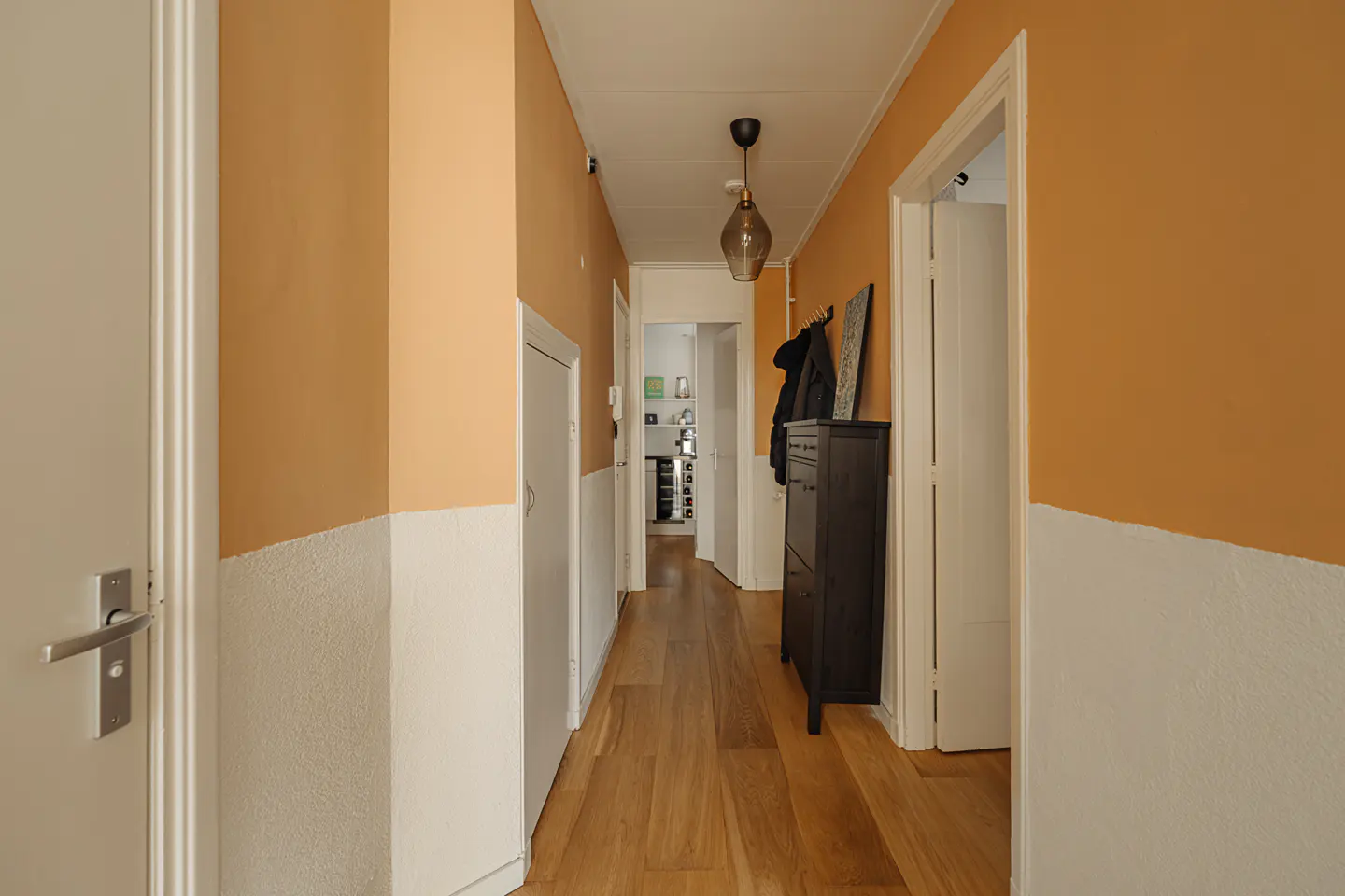 Hallway with wood floors, white doors, and walls painted white and peach. A black cabinet and coat hang near an open doorway.