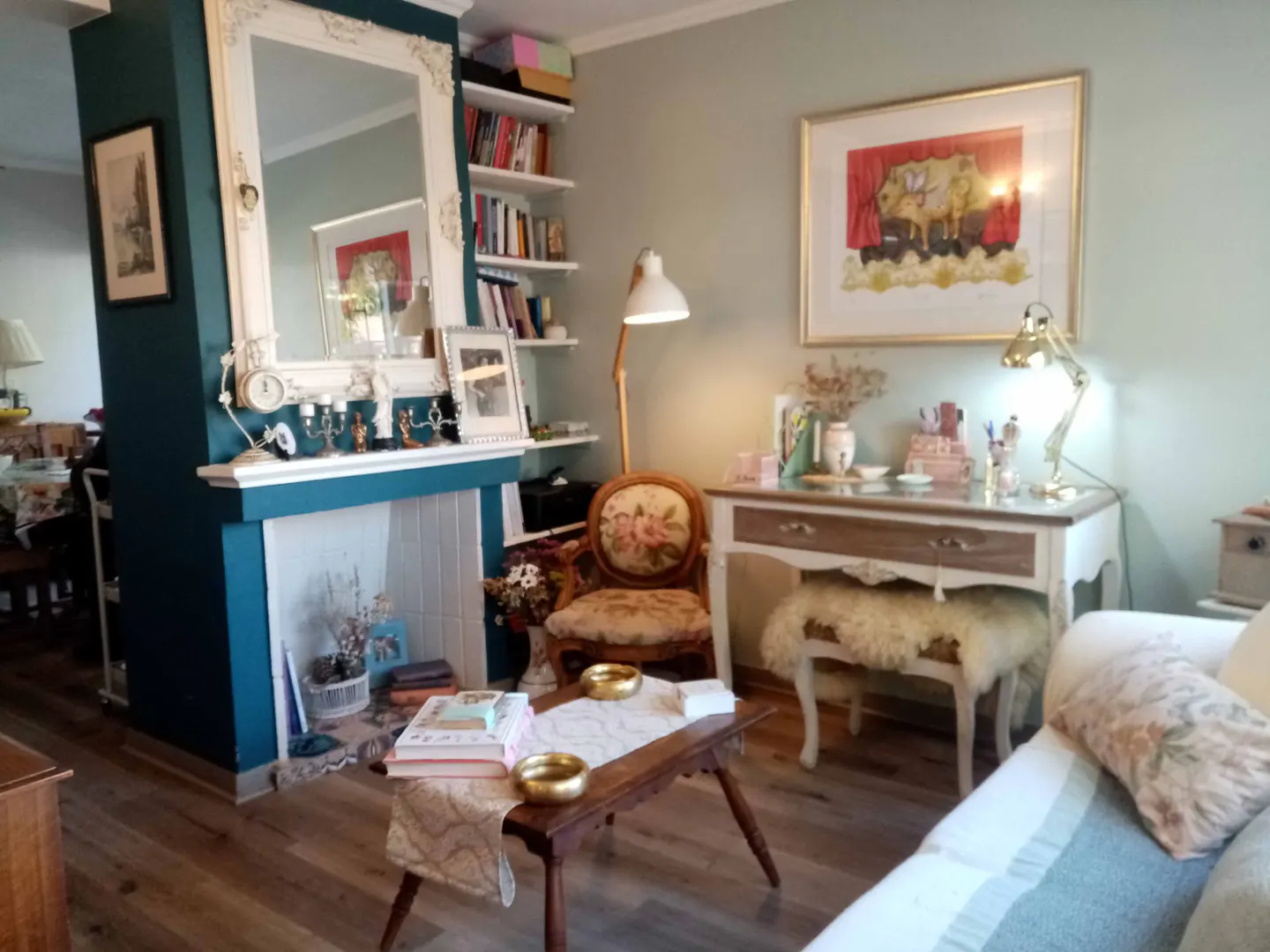 Cozy living room with a teal fireplace, white mantel, and ornate mirror. A wooden coffee table sits in front of a floral armchair and a white desk with a gold lamp.