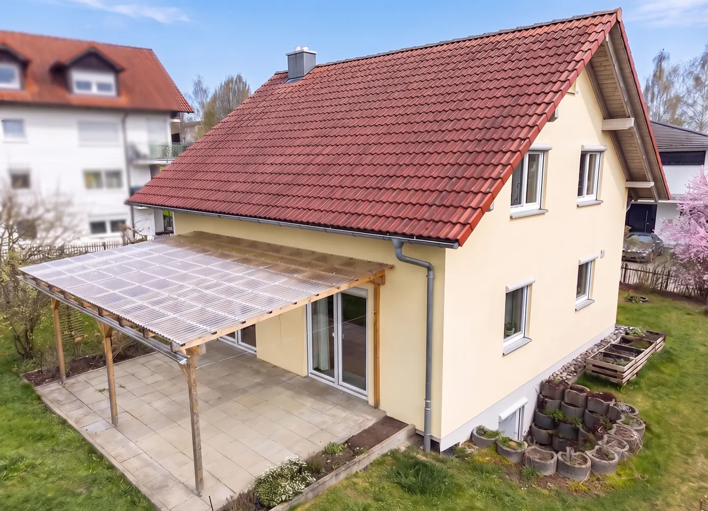 Two-story house with red tile roof and yellow siding. A patio with a clear roof is attached to the back. Green lawn and garden.