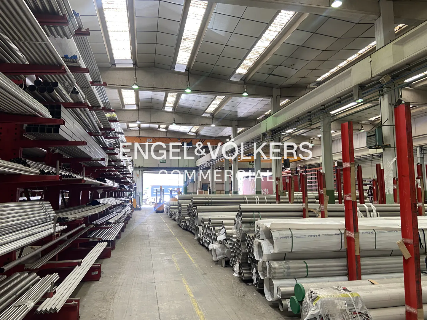 A warehouse interior with metal pipes stacked on shelves and on the floor. Overhead lighting illuminates the space.