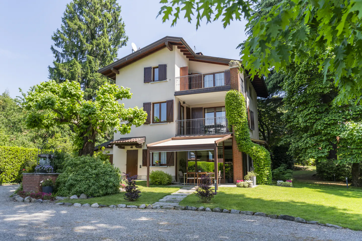Two-story house with balconies, brown shutters, and a covered patio with a table and chairs, surrounded by green trees and lawn.