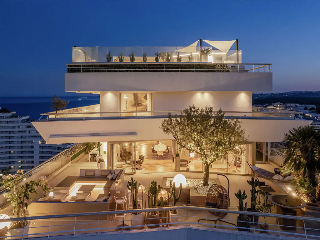 Night view of a modern white building with a large terrace featuring cacti, seating, and a tree with lights. Ocean and city lights in the background.