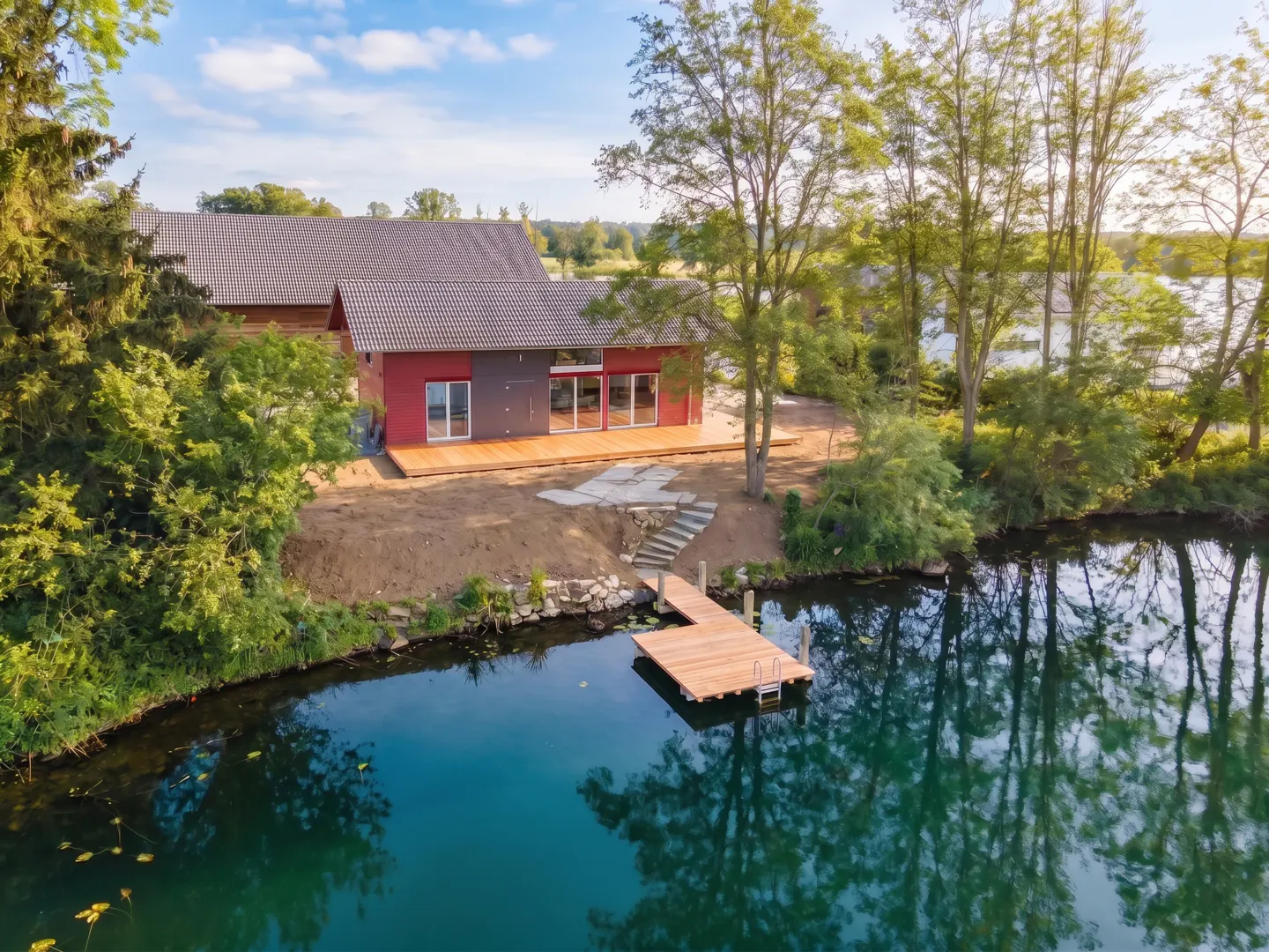 A modern red and gray house with a wooden deck and dock on a lake, surrounded by trees.