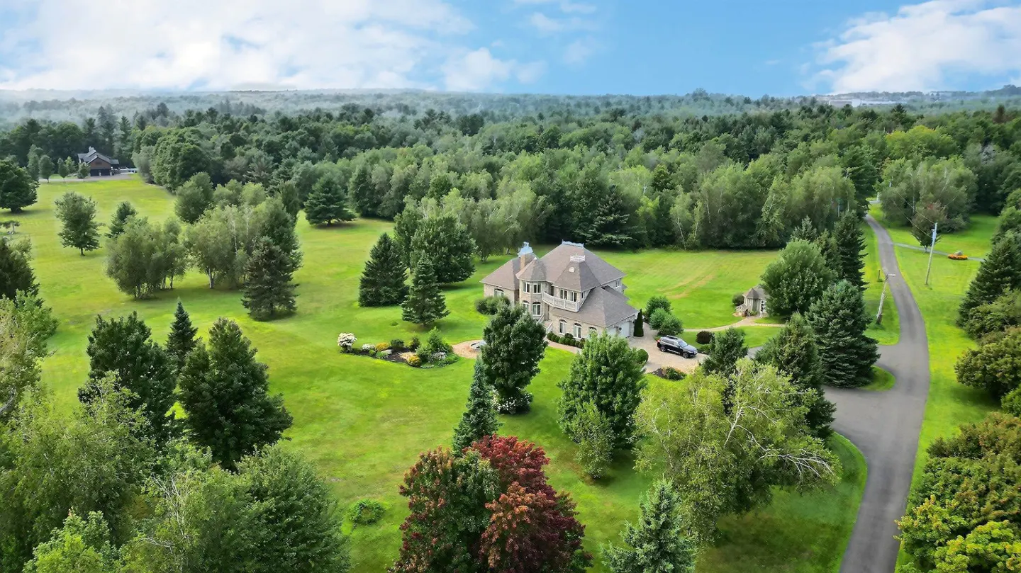 Aerial view of a large, light-colored house with a gray roof, surrounded by green lawns and trees. A driveway leads to the house.