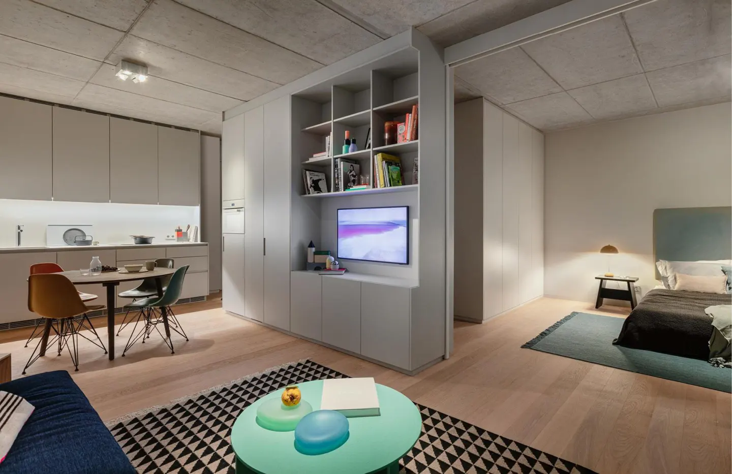 Studio apartment with a kitchen, dining area, living room, and bedroom. A light green coffee table sits on a black and white rug. A TV is mounted in a white shelving unit.
