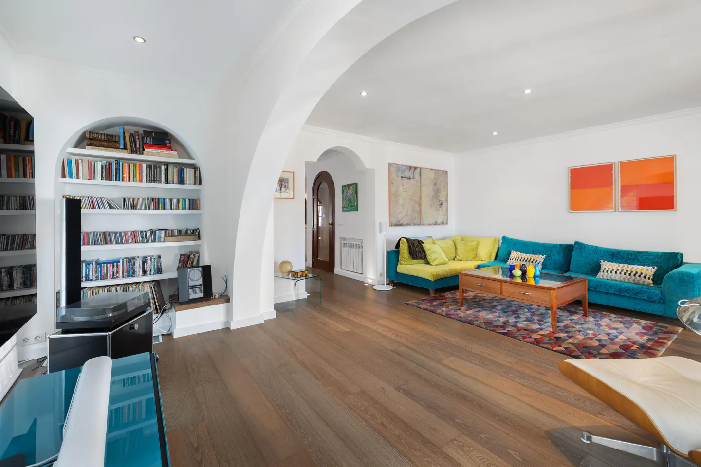 Bright living room with hardwood floors, arched built-in bookshelves, a teal sectional sofa, and colorful wall art.