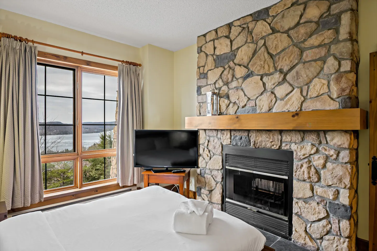 Bedroom with a stone fireplace, a TV, and a window with a view of a lake and trees. The bed has white linens.