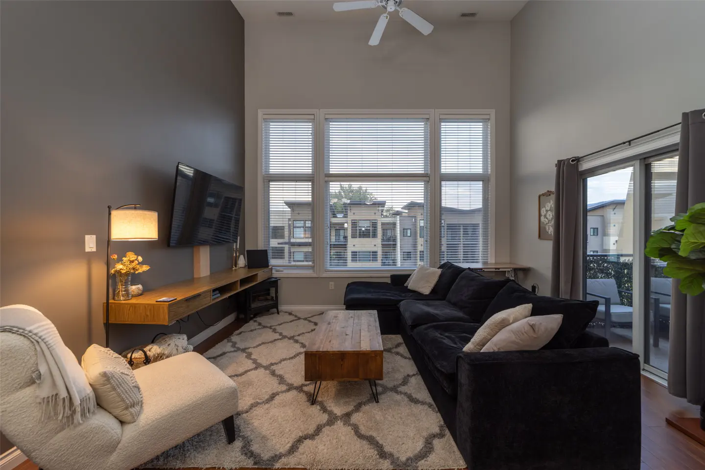 Living room with a black sectional sofa, a white chaise lounge, and a large window with blinds. A TV is mounted on the gray wall.