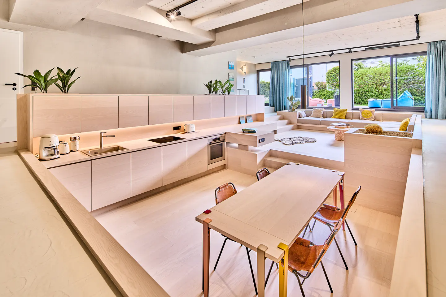 Modern open-plan kitchen and dining area with light wood cabinetry, sunken living room, and large windows with blue curtains.
