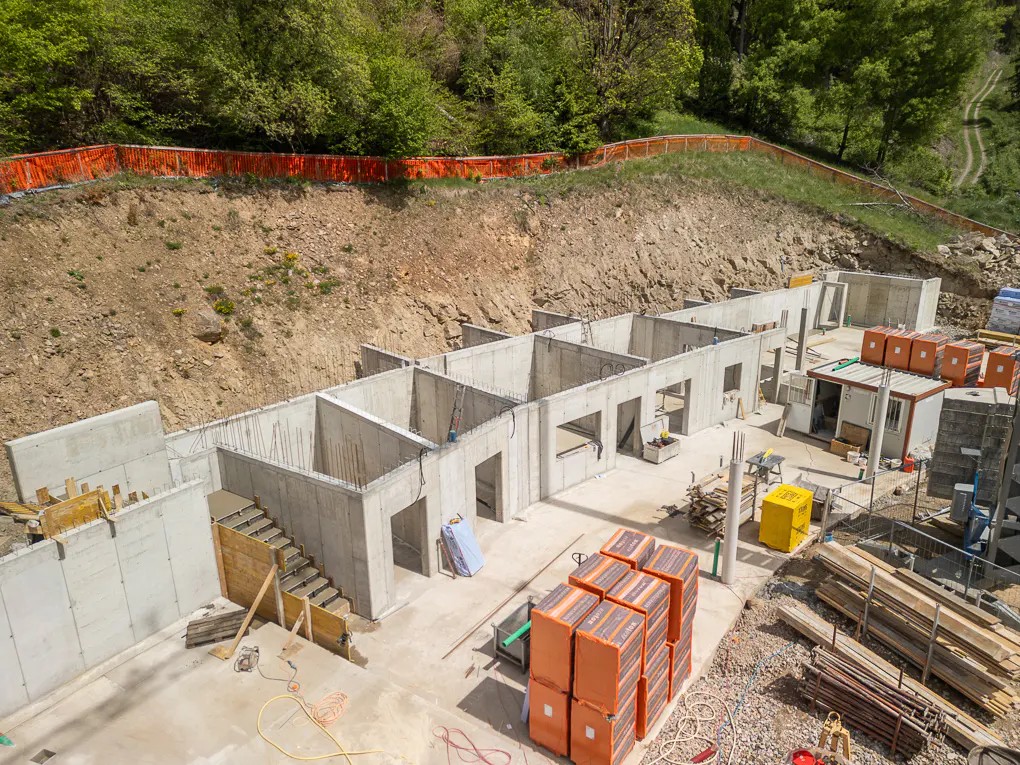 Construction site with concrete foundation walls and stairs, surrounded by dirt and trees. Orange safety fence visible.