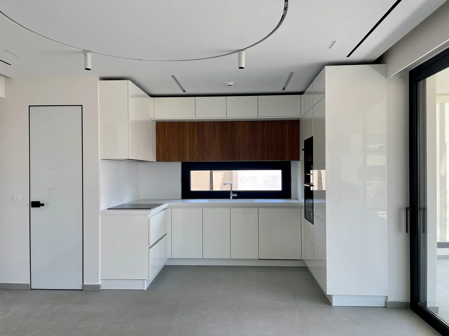 A modern kitchen with white cabinets, a wood accent wall, and a black-framed window. A white door is on the left.
