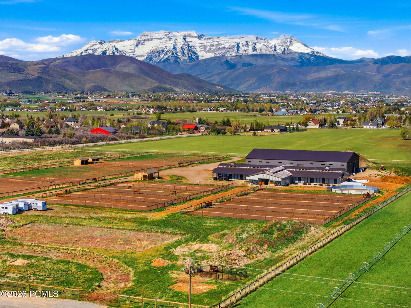 Aerial view of a horse farm with barns, paddocks, and snow-capped mountains in the background under a blue sky.