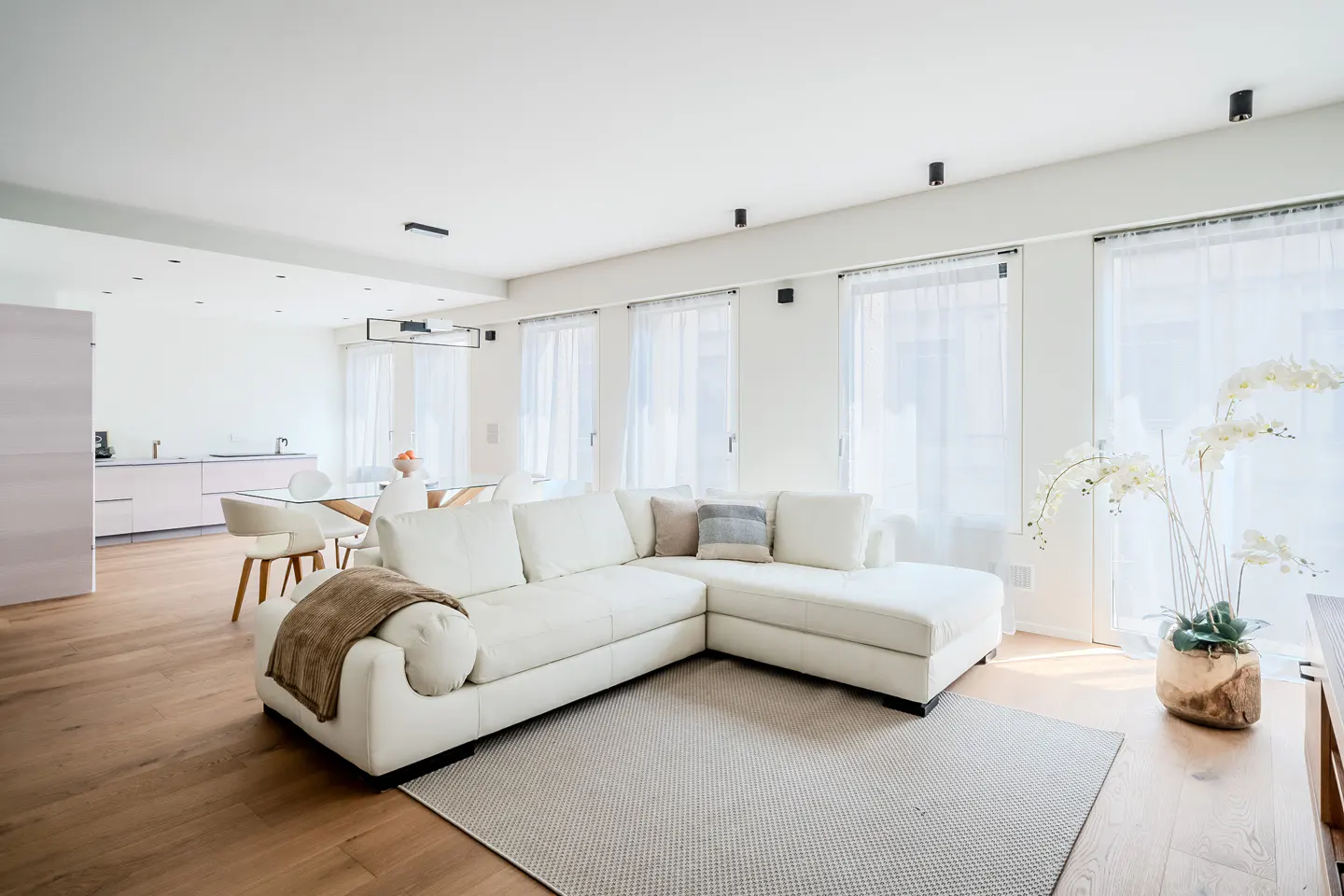 Bright, modern living room with a white sectional sofa, light wood floors, and sheer white curtains. A dining area and kitchen are visible in the background.