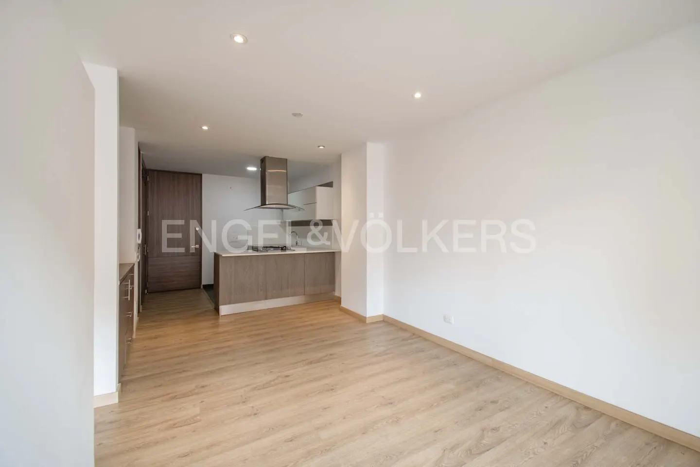 Bright, empty apartment with light wood floors and white walls. A kitchen area with gray cabinets is visible in the background.