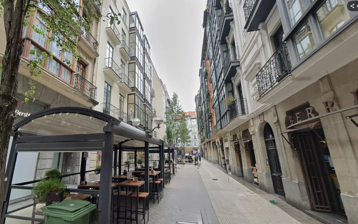 Street view of a narrow European street with outdoor cafe seating. Buildings are tall with balconies.