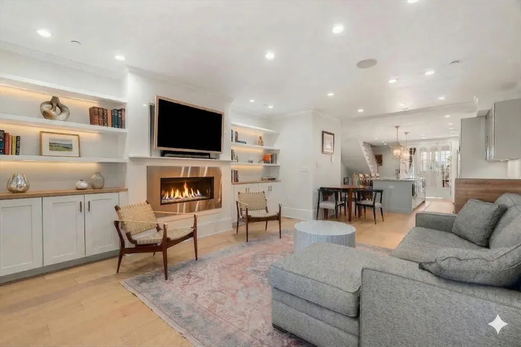 Bright, open-concept living room with a fireplace, TV, built-in shelves, and a gray sofa. A dining area and kitchen are visible in the background.