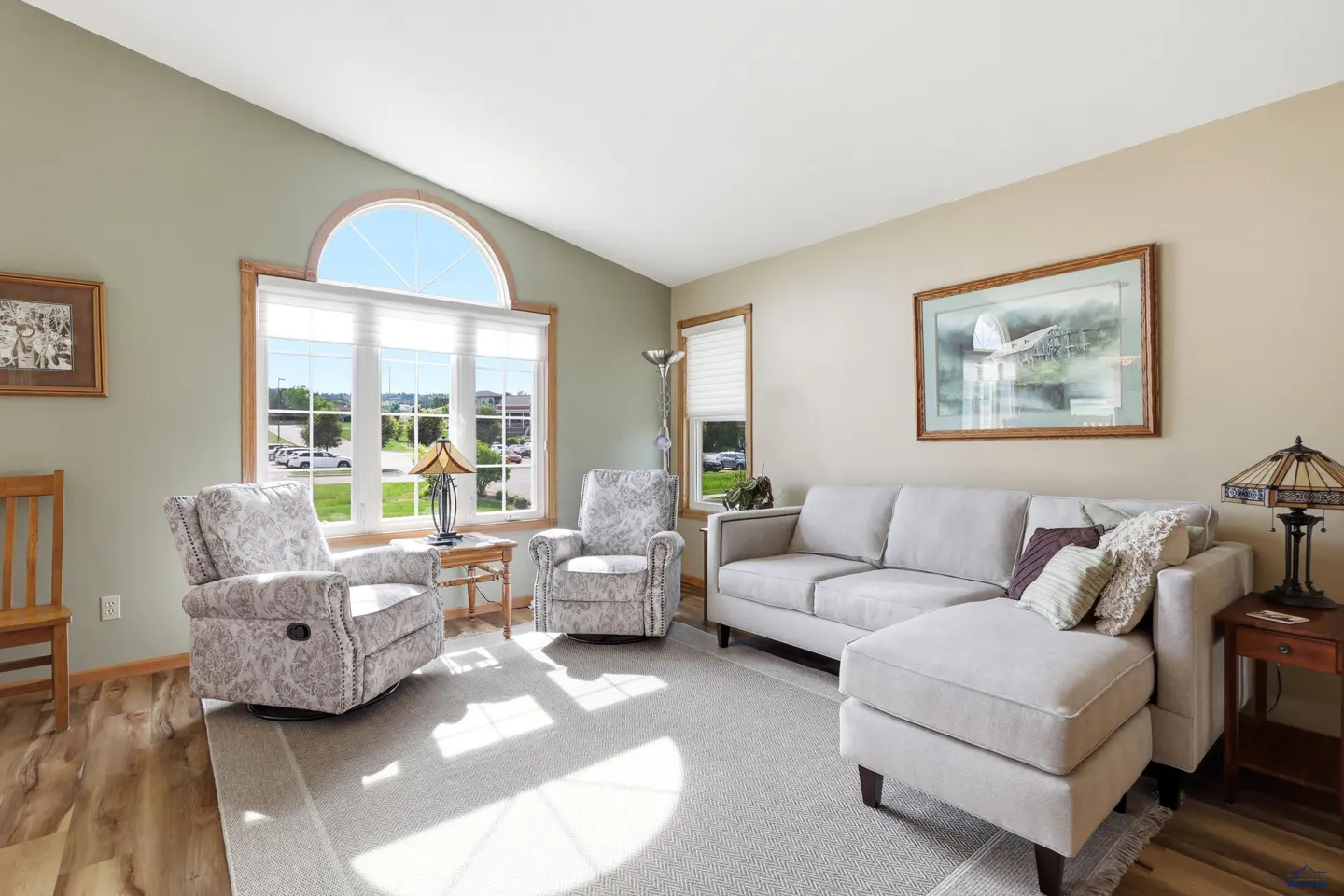 Living room with light gray sectional, two patterned armchairs, arched window, and wood-look flooring.