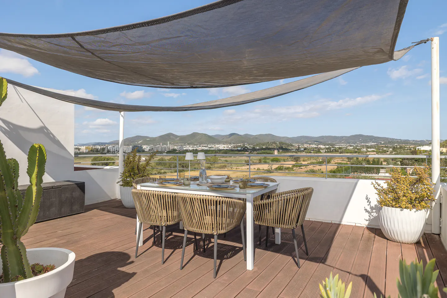 Outdoor dining area on a rooftop terrace with a table, chairs, and a sunshade, overlooking a scenic landscape.