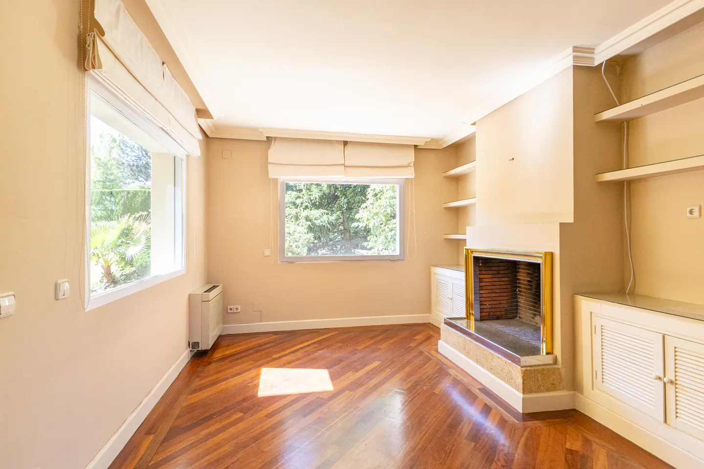 Bright living room with hardwood floors, a fireplace with gold trim, built-in shelves, and large windows with white blinds.