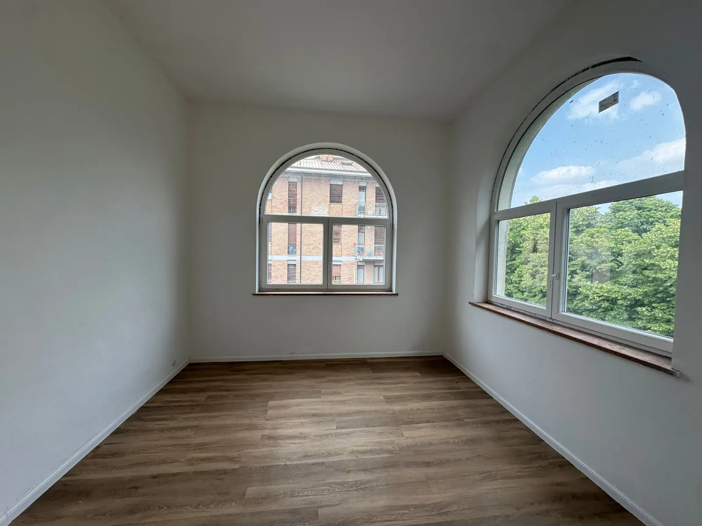 Empty room with white walls, wood-look flooring, and two large arched windows overlooking trees and a brick building.