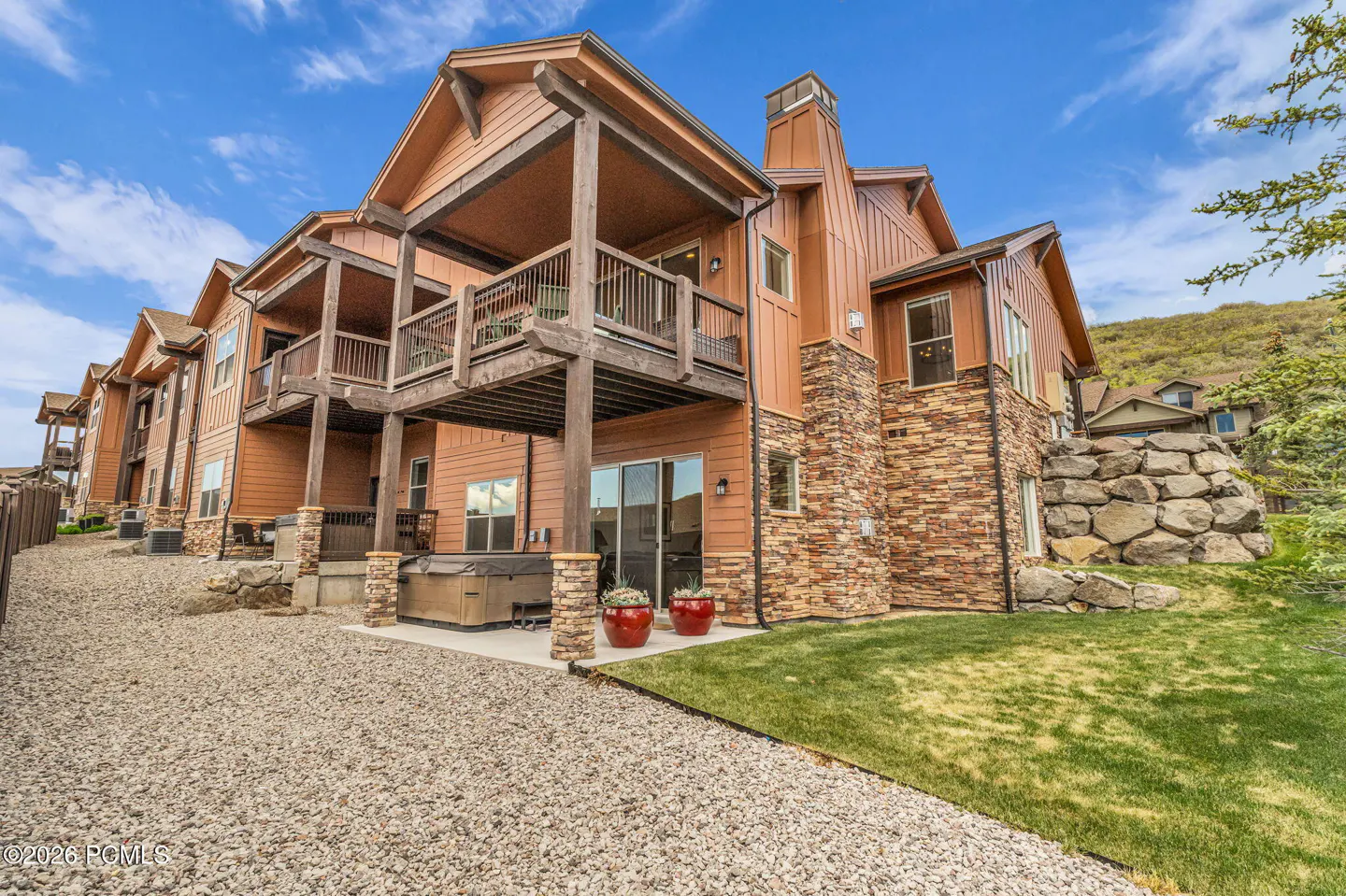 Multi-story mountain home with tan siding, stone accents, a wooden balcony, a hot tub, and a gravel yard under blue sky.