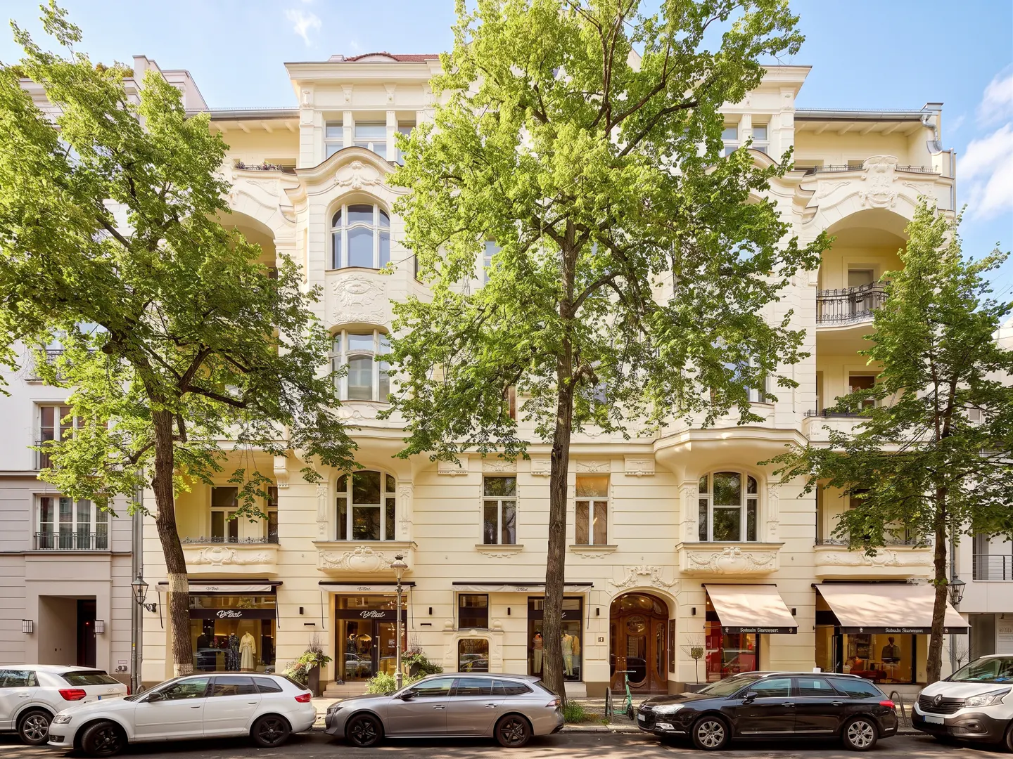 Facade of a cream-colored building with shops on the ground floor and cars parked in front.