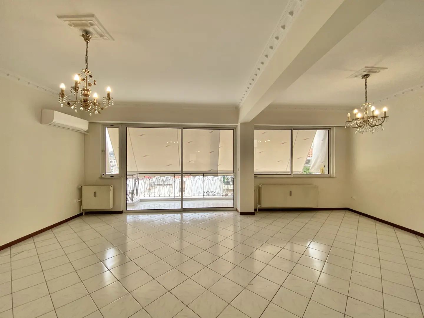 Bright, empty room with white tile floor, large windows with blinds, and two chandeliers.