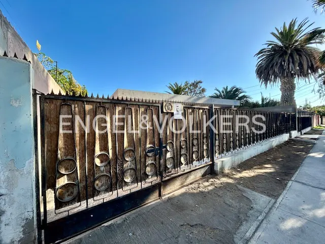 Exterior shot of a property with a wooden gate and the Engel & Volkers logo. Palm trees and a blue sky are visible in the background.