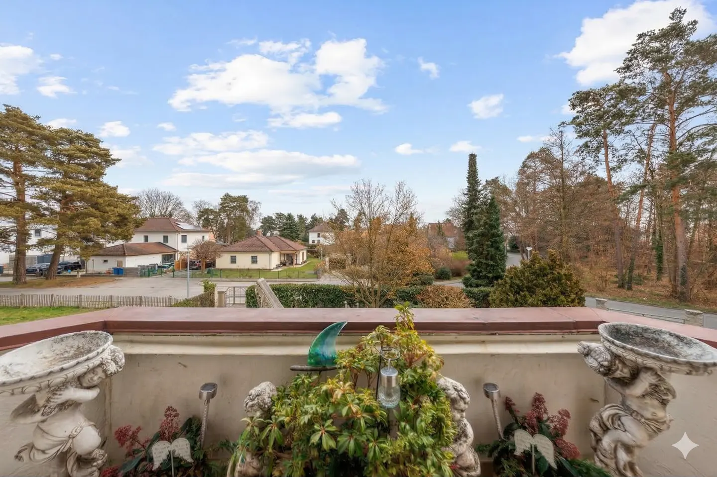 Balcony view with angel statues, plants, and a blue glass ornament. Houses and trees are visible in the background under a cloudy sky.