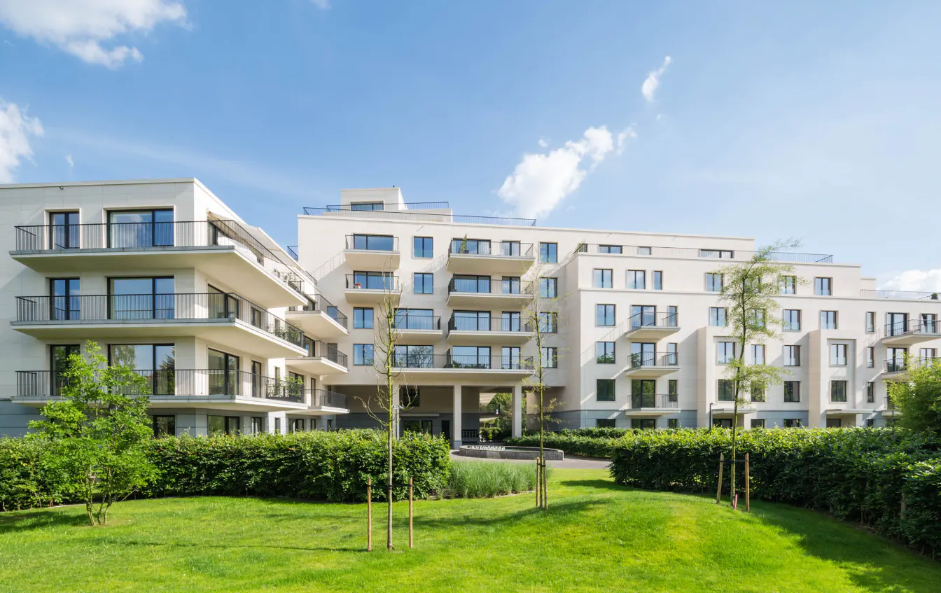 Modern, multi-story apartment building with white facade and black balconies, surrounded by green lawn and manicured hedges under a blue sky.